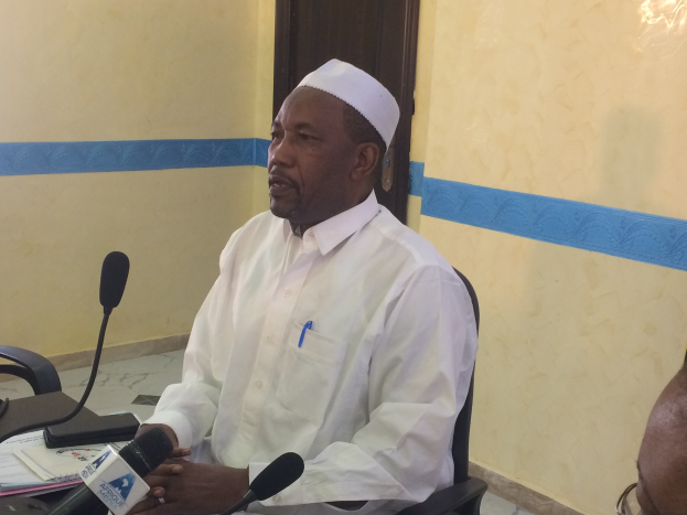 Ein Mann in einem weißen Kleid und einer Mütze sitzt an einem Tisch mit Mikrofonen und spricht, während Papiere und Gegenstände auf dem Tisch zu sehen sind; ein weiterer Mensch mit Brille ist rechts von ihm, mit einer Tür und einer Wand im Hintergrund.