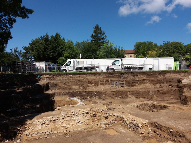 Baustelle mit einem geparkten Lkw, umgeben von einem Zaun und Bäumen, mit Baugruben im Hintergrund unter einem bewölkten Himmel.