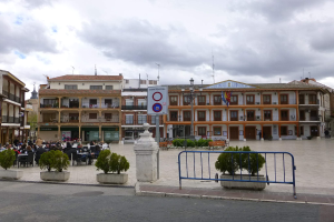 Ein belebter Stadtplatz mit Menschen, die sitzen und stehen, Topfpflanzen, Metallabsperrungen, ein Schild an einem Pfahl, Straßenlaternen mit Fahnen, umliegende Gebäude und ein bewölkter Himmel.