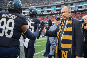Ein Mann im Anzug und Krawatte gibt einem Fußballspieler die Hand auf einem Feld, umgeben von einer Gruppe von Menschen, mit einer kameratragenden Person auf der rechten Seite und einem Stadion voller Zuschauer im Hintergrund, das Schilder trägt, die 'Notre Dame Football Coach Mike McCarthy und die Fighting Irish' lesen.
