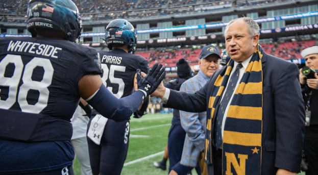 Ein Mann im Anzug und Krawatte gibt einem Fußballspieler die Hand auf einem Feld, umgeben von einer Gruppe von Menschen, mit einer kameratragenden Person auf der rechten Seite und einem Stadion voller Zuschauer im Hintergrund, das Schilder trägt, die 'Notre Dame Football Coach Mike McCarthy und die Fighting Irish' lesen.
