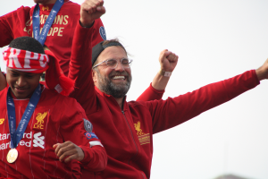 Liverpool-Trainer Jürgen Klopp feiert in roter Jacke mit seinen Spielern den Sieg der UEFA Champions League, umgeben von zwei medaillentragenden Individuen und dem Himmel im Hintergrund.