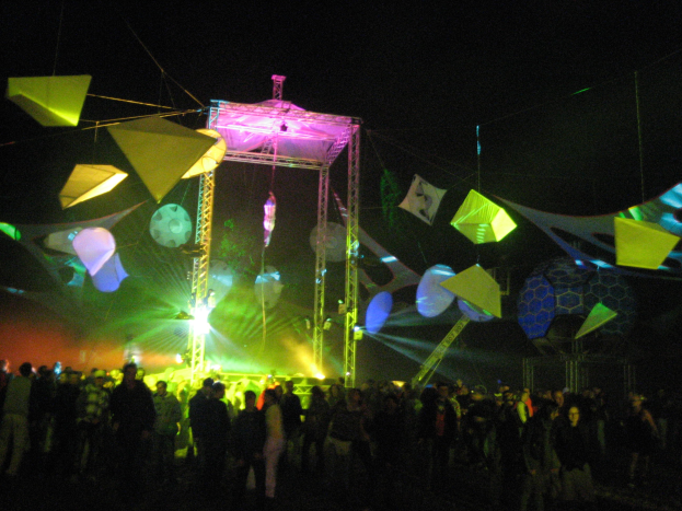 Menschenmenge vor einer hell erleuchteten Bühne bei Nacht, mit bunten Lichtern und hängenden Dekorationen vor einem dunklen Hintergrund.
