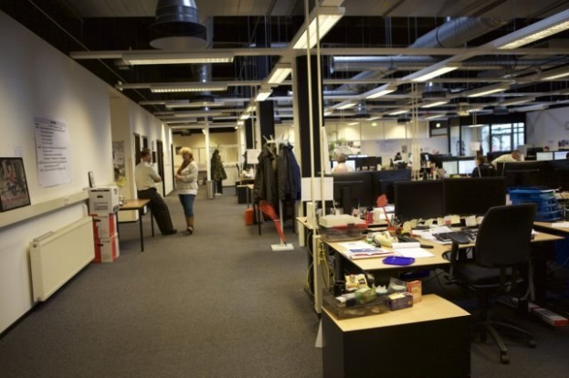 Großes Großraumbüro mit Menschen an Schreibtischen, Computern und Monitoren, umgeben von Papieren, Büchern, Kisten und Wandtafeln mit Text und Bildern unter Deckenlampen und Glasfenstern.