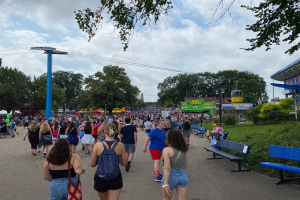 Eine Menschenmenge, die eine Straße mit Bänken, Pflanzen, Gras, Bäumen, Pfählen, Laternen, Kabeln und einem Schuppen gesäumt, mit einigen, die Taschen tragen, unter einem bewölkten Himmel.
