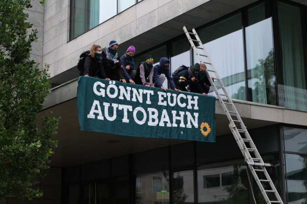 Eine Gruppe von Menschen steht auf dem Dach eines Gebäudes, hält eine Fahne, mit einem Baum links und Glasfenstern im Hintergrund, wahrscheinlich protestierend im Namen der deutschen Regierung.