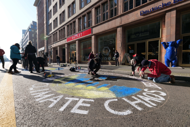 Eine Gruppe von Menschen sitzt vor einem Gebäude während einer Klimaprotest in Berlin, umgeben von Flaschen und anderen Gegenständen, mit Bäumen und einem klaren blauen Himmel im Hintergrund.