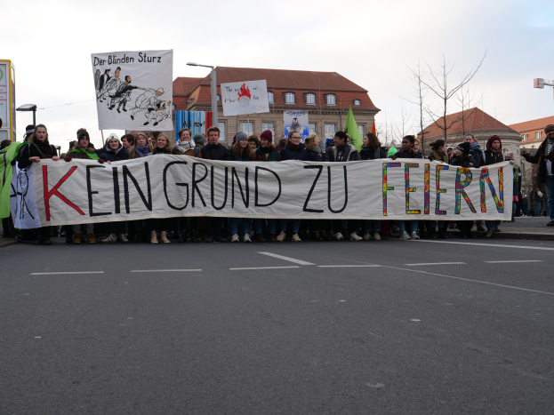 Eine Gruppe von Menschen steht auf der Straße und hält ein Schild hoch, auf dem "Kein Grund zu Feiern" steht, umgeben von Gebäuden, Laternenmasten, Bäumen und einem klaren blauen Himmel.