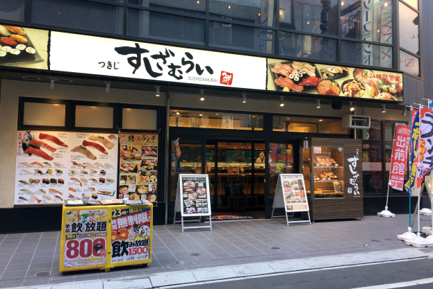 Außenansicht eines Sushi-Restaurants in Tokyo mit Glaswänden, Essensauslagen, Schildern und einer Straße davor.