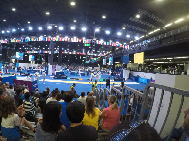 Große Menschenmenge auf Sitzplätzen und Stehplätzen in einer Indoor-Sportarena mit Bildschirmen, Flaggen und Deckenbeleuchtung.