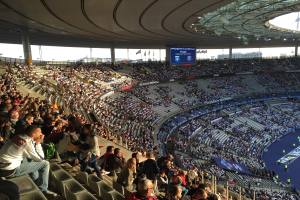 Große Menschenmenge in einem Stadion bei einem Fußballspiel mit einer Bühne, Fahnen, Stangen und einem Bildschirm im Hintergrund, identifiziert als Allianz Arena in München, Deutschland.