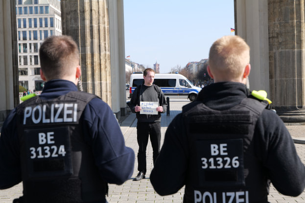 Drei Polizeibeamte stehen vor einem Gebäude, wobei ein Mann in der Mitte ein Papier hält, flankiert von Säulen; Fahrzeuge, Menschen, Bäume, Gebäude und der Himmel sind im Hintergrund sichtbar.