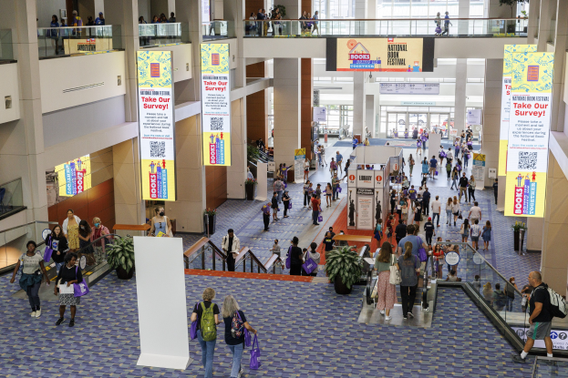 Eine große Menschenmenge geht durch ein großes Gebäude mit Bannern, Topfpflanzen, Rolltreppen, Geländern, Säulen, bedruckten Schildern, Glastüren und anderen Elementen, einige tragen Taschen, was auf den Besuch der Nationalen Buchmesse hinweist.