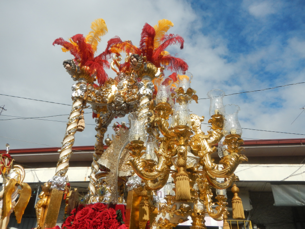 Ein prächtiger goldener und roter Festwagen, geschmückt mit Blumen und anderen Dekorationen bei einem Karnevalsumzug, mit einem Gebäude, Strommasten mit Kabeln und einem bewölkten Himmel im Hintergrund.