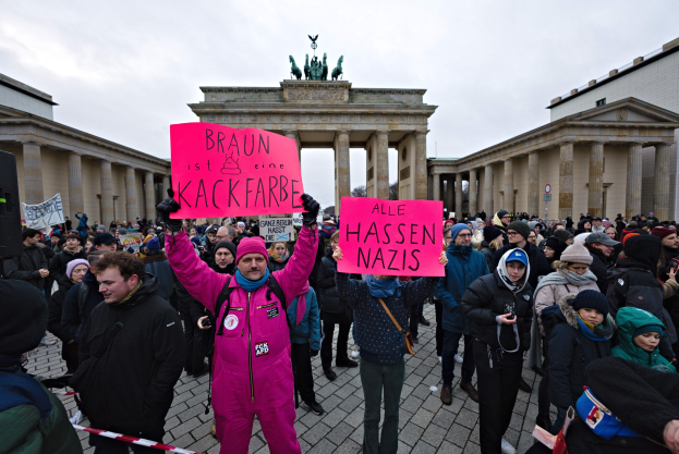 Gruppe von Menschen protestiert vor dem Reichstagsgebäude in Berlin, hält Schilder gegen die Anti-Nazi-Bewegung der deutschen Regierung, mit Gebäuden, Säulen und Statuen im Hintergrund unter einem klaren blauen Himmel.
