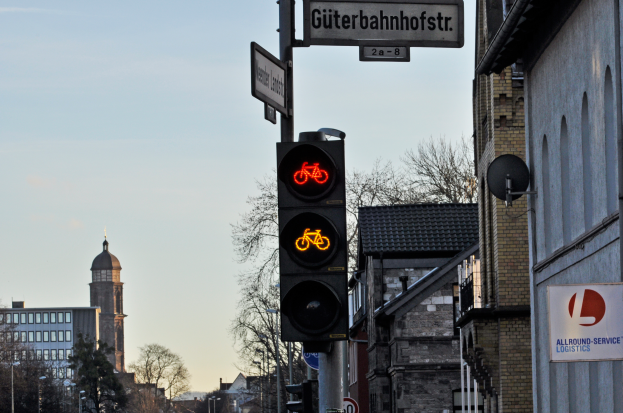 Eine Ampel mit einem Fahrradschild in einer Stadt, umgeben von Gebäuden, Bäumen, Pfosten, Schildern und Fahrzeugen auf der Straße, mit dem Himmel im Hintergrund.