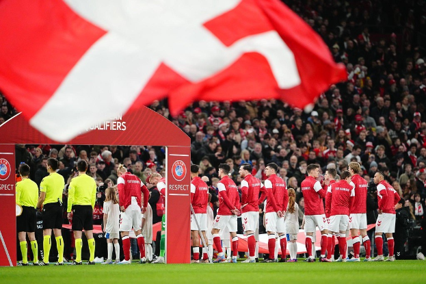 Gruppe von Menschen auf einem Fußballfeld mit einer roten und weißen Flagge im Vordergrund, einem Bogen mit der Aufschrift "Bayern München vs. Bayern München Wett-Tipps & Vorschau" im Hintergrund und einer großen Menschenmenge im Stadion.