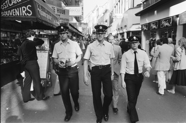 Drei Polizisten in Uniform gehen eine belebte Straße mit hohen Gebäuden und Geschäften entlang, mit einem klaren Himmel im Hintergrund.
