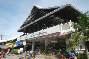 Outdoor-Markt in Koh Samui, Thailand, mit Menschen, geparkten Fahrzeugen, einem Metallzaun, Bäumen, einem Banner und einem Dach mit Deckenleuchten unter einem bewölktem Himmel.