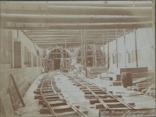 Schwarzes und weißes Foto einer Eisenbahnschiene in einem Gebäude unter Bau, mit verstreuten hölzernen Gegenständen, Fässern, Wänden, Pfählen und Text am unteren Rand.