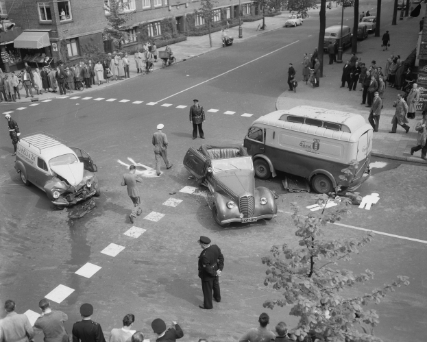 Schwarz-weiß-Szene eines Autounfalls an einer Straßenkreuzung mit mehreren Fahrzeugen, darunter ein Van, und einer Gruppe von Menschen, die danebenstehen, mit Gebäuden, Bäumen, Polen und Texttafeln im Hintergrund.