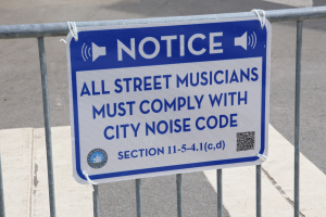 Anschlagtafel an einem Zaun mit einem Schild, auf dem "Anschlag: Alle Straßenmusikanten müssen die Stadtlärmordnung einhalten" steht und eine Straße im Hintergrund.