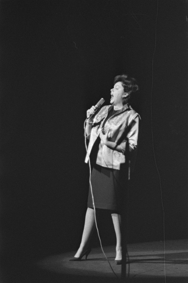 Eine Frau in einem schwarzen und weißen Outfit mit 1950er-Jahre-Frisur singt fröhlich auf einer von einem Scheinwerfer beleuchteten Bühne in ein Mikrofon.