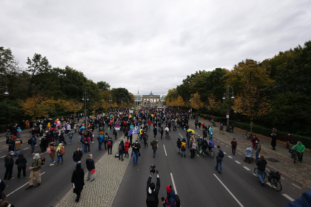 Eine große Gruppe von Menschen geht eine von Bäumen gesäumte Straße mit Laternenpfählen entlang, hält Kameras in der Hand, während sie an einer Protestdemo in Berlin teilnehmen, mit einem Gebäude und einem klaren Himmel im Hintergrund.