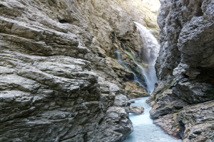 Ein kleiner Wasserfall stürzt eine steile Felsenschlucht hinab, umgeben von grünen Hügeln, mit klarem Wasser, zerklüfteten Felsen und hellem Sonnenlicht, das die Szene erhellt.