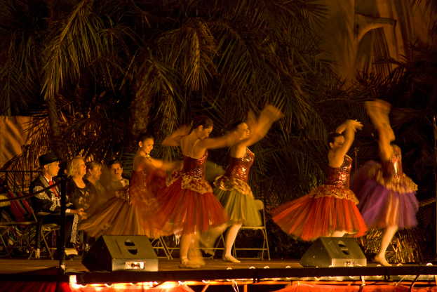 Leute tanzen auf einer Bühne mit Scheinwerfern und Bäumen im Hintergrund, während andere auf Stühlen im Dunkeln sitzen.