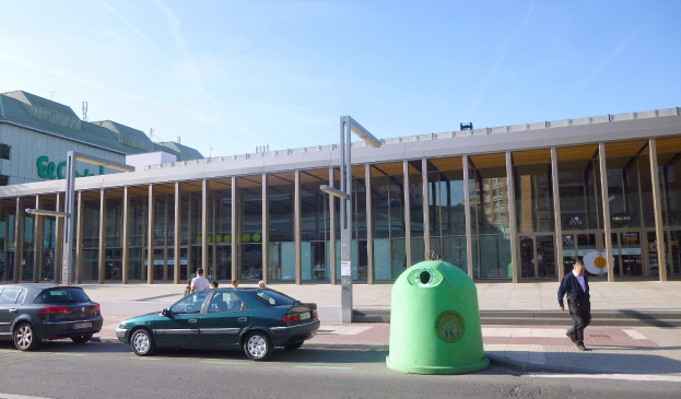 Grüner Müllcontainer auf einer Stadtstraße neben einem Glaswandgebäude mit Säulen, mit Fahrzeugen und Passanten in der Nähe, unter einem bewölkten Himmel am Berliner Hauptbahnhof.