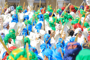 Eine große Gruppe von Menschen in bunten Kostümen marschiert eine Straße entlang, jeder hält einen Stock in der Hand, mit einem Gebäude im Hintergrund.