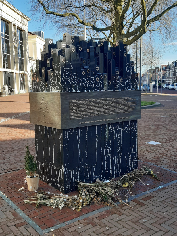 Denkmal für Holocaust-Opfer in einem Stadtplatz mit einem Baum davor, Topfpflanze und trockenen Blättern auf dem Boden, umgeben von Gebäuden, Fahrzeugen, Masten und Bäumen unter dem Himmel.