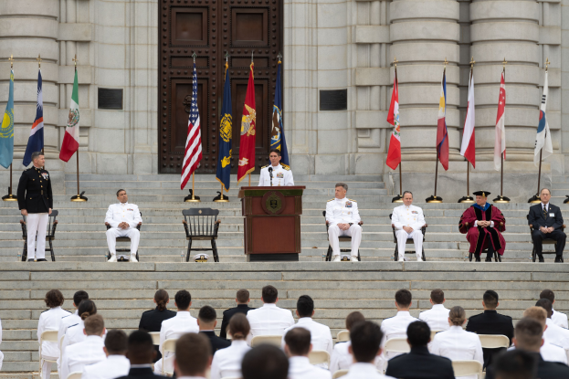 Gruppe von Menschen in weißen Uniformen auf Treppe eines Gebäudes mit Podium und Flaggen vor großem Gebäude mit Tür während einer Ehrenzeremonie.