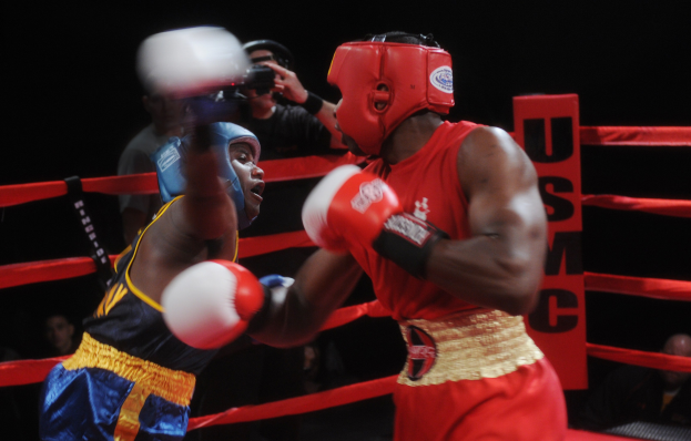 Zwei Boxer im Ring während eines Kampfes, mit einem Fotografen und Zuschauern im Hintergrund bei gedämpftem Licht.