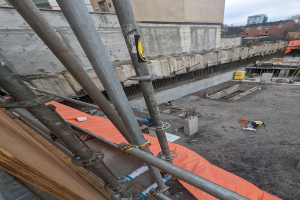 Eine Baustelle mit einem großen Gebäude im Hintergrund, umgeben von Bäumen und einem klaren blauen Himmel, mit Metallstangen, Rohren und Arbeitern im Vordergrund.