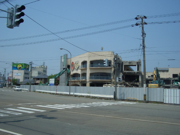 Stadtstraße mit Fahrzeugen, Strommasten, Laternen, einer Ampel auf der linken Seite, Gebäuden und Bäumen im Hintergrund und einem teilweise abgerissenen Gebäude im Vordergrund.