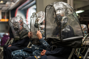 Eine Gruppe von Menschen in Schutzausrüstung sitzt auf einem Zug, mit Gegenständen auf der rechten Seite und Lichtern an der Spitze des Daches, vor einem unscharfen Hintergrund.
