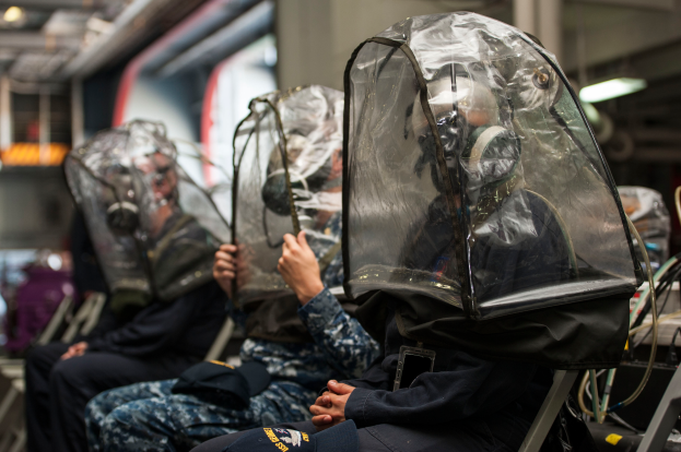 Eine Gruppe von Menschen in Schutzausrüstung sitzt auf einem Zug, mit Gegenständen auf der rechten Seite und Lichtern an der Spitze des Daches, vor einem unscharfen Hintergrund.