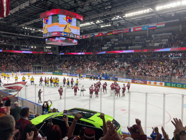 Ein Eishockeyspiel in einer großen Arena mit Zuschauern, ein Auto vor dem Gebäude, eine von einem Zaun umgebene Eisfläche, Banner mit Text und Deckenbeleuchtung und Bildschirm mit hängenden Fahnen.