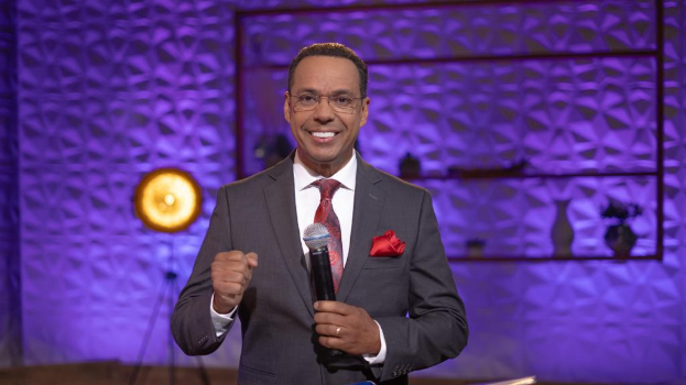 Stephen Colbert in Anzug, Krawatte und Brille auf einer B├╝hne stehend und ein Mikrofon haltend, mit einer Wand und verschiedenen Gegenst├Ąnden dahinter.