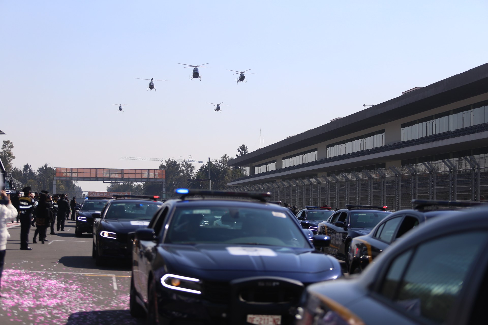 Polizeifahrzeuge vor einem Gebäude mit Menschen auf der linken Seite, Hubschrauber am Himmel und eine Brücke im Hintergrund.