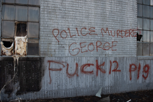 Ein Gebäude mit Graffiti, das "Polizei ermordet George Fuck 12 P19" auf der Seite trägt, mit Fenstern und verschiedenen Gegenständen vor ihm.