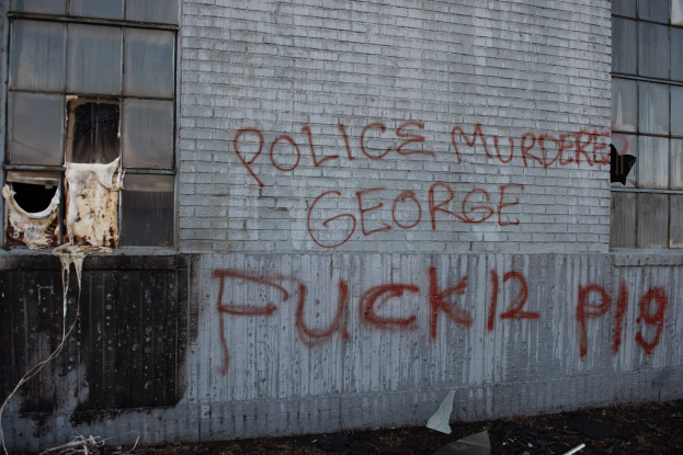 Ein Gebäude mit Graffiti, das "Polizei ermordet George Fuck 12 P19" auf der Seite trägt, mit Fenstern und verschiedenen Gegenständen vor ihm.