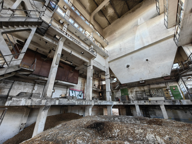 Verlassene Fabrik im Innern mit einem großen Haufen Erde im Vordergrund, mit Treppen, Geländern, Säulen, Wänden, Fenstern, Lampen und einem Sandhaufen am Boden.