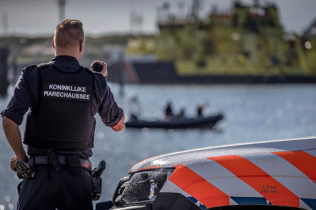 Ein Polizist in Uniform steht neben einem Polizeiwagen an einem Gewässer und hält ein Funkgerät in der Hand. Im Hintergrund ist ein Boot mit ein paar Menschen auf dem Wasser zu sehen.