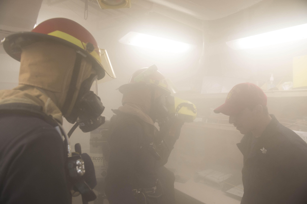 Feuerwehrleute in Helmen und Ausrüstung atmen Rauch während einer Übungseinheit aus, wobei einer eine Kamera und Ausrüstung im Hintergrund hält.