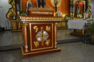 Ein Altar in einer Kirche mit einem Kreuz, Statuen, Blumensträußen, Kerzen, einem Tisch mit einer Decke und anderen Gegenständen, umgeben von Wänden, die mit Lichtern geschmückt sind.