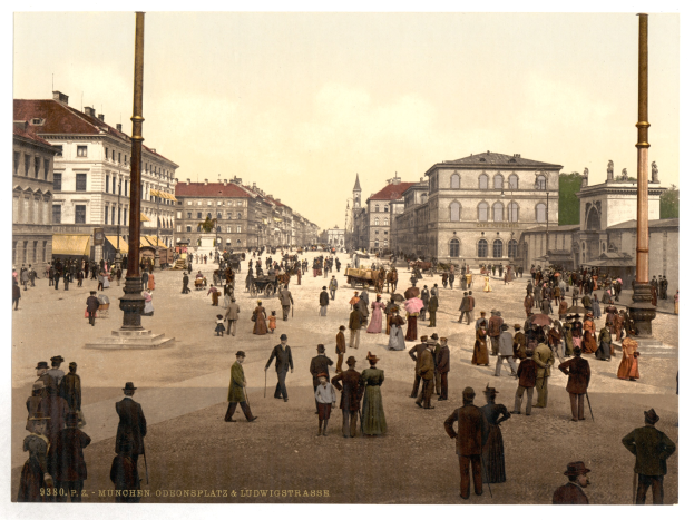 Schwarzes und weißes Foto einer belebten Münchner Straße in den frühen 1900er Jahren mit Fußgängern, einem Pferdewagen, Gebäuden, Bäumen und einem sichtbaren Himmel.