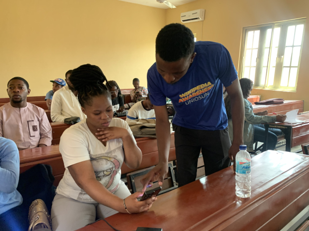 Eine Gruppe von Menschen sitzt an Tischen in einem Klassenzimmer, wobei ein Mann steht und ein Mobiltelefon hält, Flaschen und andere Gegenstände auf den Tischen, eine Klimaanlage an der Wand und ein Fenster im Hintergrund.